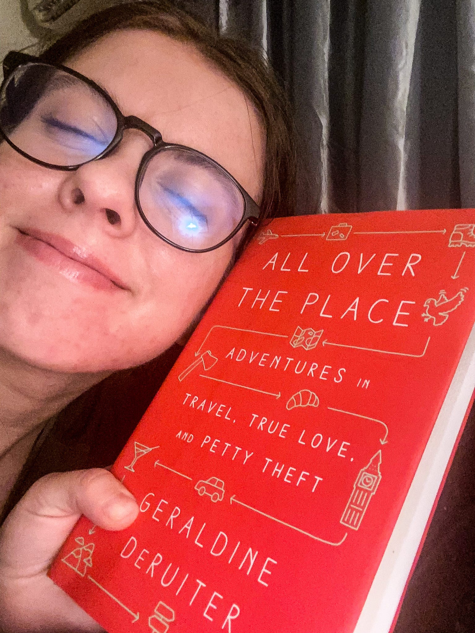 A photo of me smiling like a dork in my blue light glasses while hugging "All Over the Place" - a red book by Geraldine DeRuiter