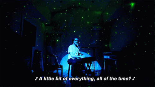 A gif of a white man in round sunglasses playing on a keyboard with green lights all over his room.