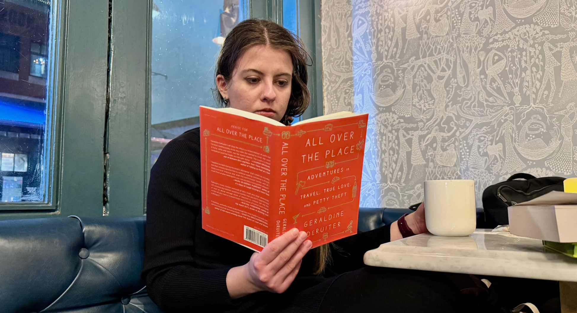 A photo of a white woman, Mariya Delano, sitting in a cafe holding a copy of "All Over The Place" by Geraldine DeRuiter.