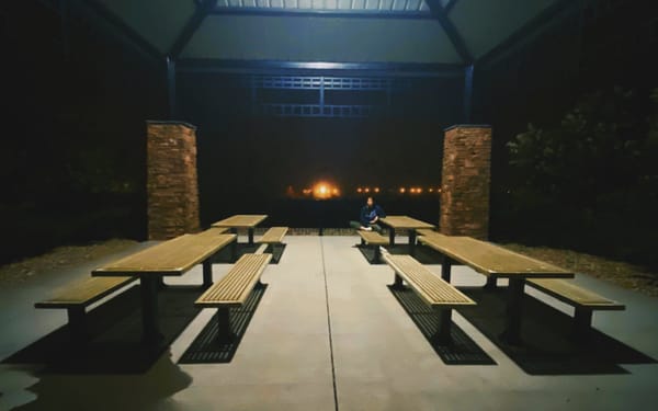 A photo of Mariya Delano sitting in a dark and empty gazebo, on a bench furthest from the camera.